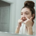 A young woman applies skincare in front of a bathroom mirror, focusing on facial care.